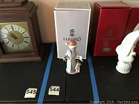 Lladro figurine with original packaging placed next to a clock and another figurine, showing the entire figure and its box.