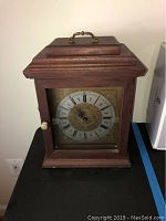 Closed view of rectangular wooden quartz mantel clock with brass handle and detailed clock face with Roman numerals.
