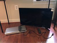 Samsung TV with screen off, remote on floor next to Toshiba DVD player, and power cords visible on hardwood floor.