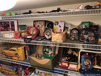 Wide view of shelves showing boxed board games Life and Mouse Trap, multiple toy cars, baskets with toy parts, and boxed NASCAR Barbie doll on the top shelf. Multiple model airplanes lined up on the top of the shelf.