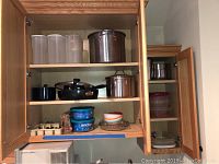 Kitchen cabinet with multiple pots, pans, food storage containers, flatware visible inside wooden cabinet shelves.