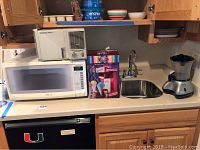 Countertop with Panasonic microwave, boxed Nostalgia Electrics 50s Style Snow Cone Maker, Kenmore air purifier on top of microwave, and Back to Basics Cocoa Latte Machine next to sink.
