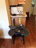 Photo showing Guitar Hero drum set with drum pads and bass pedal, two Guitar Hero guitar controllers, and a microphone, all positioned on and around a counter.