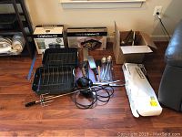 Full view of lot showing unopened boxed Calphalon stock pot and grill pan box, two roasters, electric rotisserie parts and uninstalled metal pot rack.