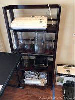 Full view of dark brown wooden shelving unit holding white FoodSaver V825 vacuum sealer, clear plastic containers of various sizes, and vacuum sealer accessories including rolls and hoses.