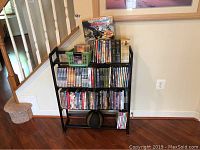 Black metal storage rack against wall near stairs with multiple DVDs and a green plastic basket with CDs on top. DVDs include various titles stacked on three shelves.
