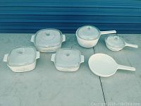 Full set overview of 11 vintage Corningware cookware pieces with clear Pyrex lids, showing various square and round dishes, saucepans, and skillets with floral patterns.