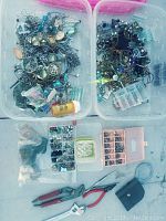 Two plastic bins filled with mixed jewelry parts including chains, charms, beads, and earrings. Several small plastic containers with beads and findings, along with jewelry pliers and a pill bottle for storage visible.