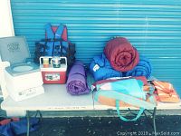 All camping items including sleeping bags, rolled tents, camping toilet, cooler, lantern, life jacket and thermos cups displayed on a table against a blue garage door.