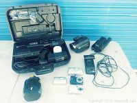 Wide shot of all 3 video camcorders, 2 digital cameras, 1 vintage camera, camera case, and cables on white surface.