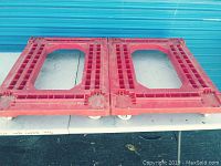 Two red four-wheel furniture dollies side by side on a table against a blue background. The dollies are rectangular with a central open cut-out and grid pattern.