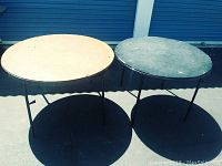 Two round card tables, side by side, one lighter and one darker, both with black metal legs, outdoors in sunlight showing overall wear and surface marks.