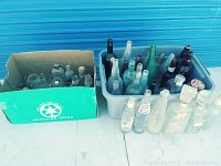 Wide-angle photo showing the large assortment of vintage bottles in a cardboard box and plastic bin. Bottles vary in size and color, predominantly clear glass with some green.