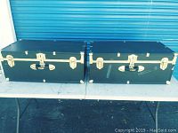 Two black Seward foot lockers placed side by side with metal hardware and minor wear.