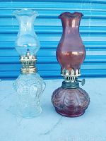 Two vintage oil lamps on white surface with blue background, one clear glass and one purple glass.