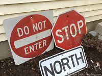 Photo showing three metal road signs: square DO NOT ENTER, octagonal STOP, and rectangular NORTH signs leaning against a wall, outdoors on soil.