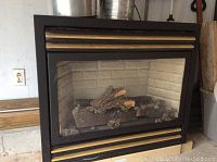 Front view of gas fireplace insert showing black metal frame, decorative gold bars, glass front, and ceramic log set inside against light colored brick patterned back panel.