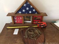 Wide photo showing the memorial display case with the folded American flag and pins alongside the wooden Navy plaque on a table with measuring tape and label showing lot number.
