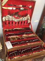 Complete flatware set displayed in open red velvet-lined wooden box, showing knives, forks, spoons, and serving utensils organized inside.