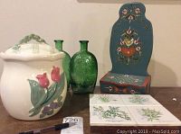 Vintage ceramic cookie jar with raised tulip floral design, two green embossed glass bottles, botanical tile trivet, and blue painted wooden storage box on a table.