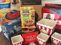 Photo showing several items including Pampered Chef loaf pans box, Perfect Tortilla pan set box, Onion Baker boxed item, popcorn-themed stoneware bakeware, and a can duster