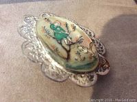 Close-up photo of the mother of pearl pin showing hand painted figure in green attire and a dog against a tree background on the oval pearl. Silver-tone filigree metal backing.