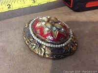 Close up side angle view of the round red and gold brooch showing detailed pearl and rhinestone decoration with measuring tape for scale.