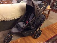 Side view of brown and grey jogging stroller with front and rear seat, canopy, three wheels on wooden floor near couch