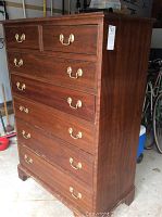 Overall view of the high chest dresser showing front and one side with brass handles on drawers. Surface wear visible at base.