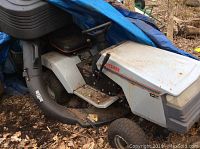 Photo showing side profile of the white Craftsman 12 HP mower with grass/leaf collection chute and black seat, stored outdoors with signs of surface dirt and rust.