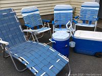 View of three blue and white vinyl folding chairs, one vintage reclining lounge chair with blue vinyl webbing, round cooler, and large cooler with wheels set against house siding.