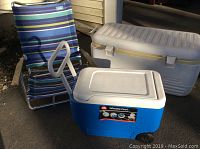 Photo showing two Igloo coolers, one large off-white cooler with more scratches and an older look, and a smaller blue wheeled cooler, alongside a blue and green striped beach chair.