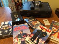 Sony VHS player with remote control and 10 VHS tapes placed on a wooden table. VHS tape titles visible include Mission Impossible 2, Casablanca, Rush Hour, and others.