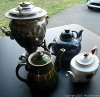 Full lot showing Russian electric urn and three distinct ceramic teapots arranged on black tabletop outdoors