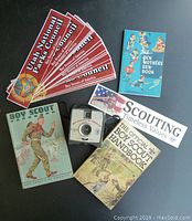 Overview photo showing the 20 bumper stickers, three books including The Den Mothers Book, Boy Scout Handbook, Official Boy Scout Handbook, and the Official Camera #127