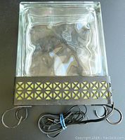 Top view of a rectangular clear glass block with wavy internal pattern, mounted in a metal frame featuring geometric cutouts. Showing attached electrical cord coiled beneath and metal hooks on each side.