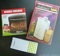 Photo of unopened George Foreman 2 Serving Grill box, Hamilton Beach HealthSmart Juice Extractor box, and Sharper Image slicer/cutter on a dark surface.