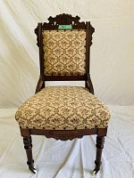 Front view of antique wooden chair showing ornate carved top rail, upholstered back and seat with floral patterned fabric, and turned front legs with casters.