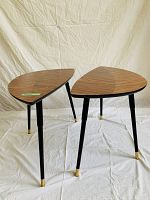 Two leaf-shaped tables on a white cloth background showing the wood grain laminate tops and black legs with gold tips.