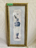 Photo shows framed vertical artwork of a blue vase with a white flower, enclosed in a gold, ornate frame.