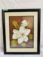 Framed art showing a white magnolia flower with green leaves on a reddish brown background, signed by Vivian Flash. Frame is black and decorative with cream matting.