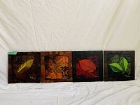Four square canvas wall art pieces each displaying a different leaf on varied artistic backgrounds laid out side by side against a white cloth background.