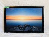 Framed photographic print showing sunset over calm water and dark rocks, with black modern frame, laid against white fabric background.