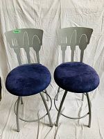 Two metal bar stools with silver frames and blue fabric cushions. The backs have cutlery silhouettes (fork, knife, spoon) cut out of metal.