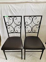 Two black metal kitchen chairs with scrollwork backs and black cushioned seats side by side, shown from the front.