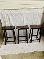 Three bar stools arranged side by side against a white sheet background on concrete floor. The stools have dark brown rectangular wooden seats and are supported with crossbar bracing.