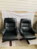 Pair of black vinyl padded lounge chairs with high backs, cushioned armrests, and wooden swivel bases shown from front view against a white backdrop.