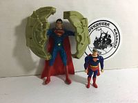 Two Superman action figures shown. Larger figure has a detachable circular shield split into two halves with a mechanical design. Both figures wear classic costume with red capes and blue suits.