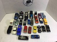 Top-down view of assortment of 24 toy cars, trucks, and buses in various colors and styles, arranged in rows on a white surface with backdrop.