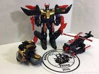 Photo showing three transforming robot toys: one standing robot in black, red, purple, and gold; one helmet-shaped vehicle in black and gold; and one black and red helicopter vehicle.